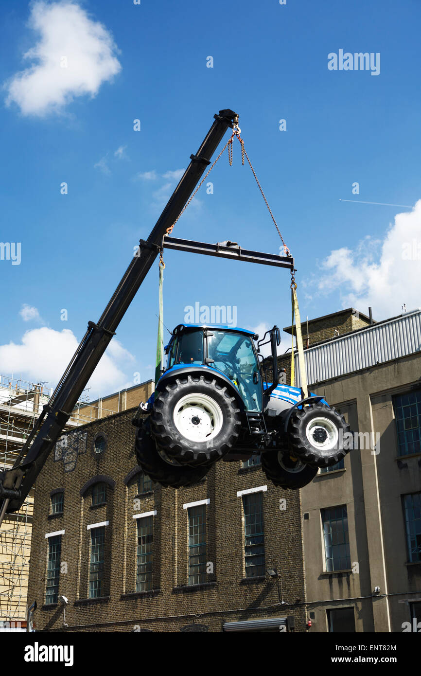 Trattore sospeso dalla gru, magazzini sullo sfondo, cielo blu chiaro. (copertina libro). Concetto di gravità. Trattore New Holland. Foto Stock