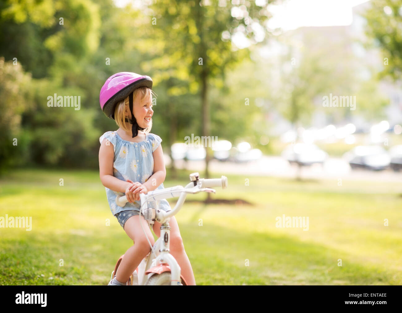 Una giovane ragazza si siede sulla sua bicicletta in un parco cittadino, sorridente, riposo, e felice. Indossa un casco rosa, e sa come mettere in pratica b Foto Stock