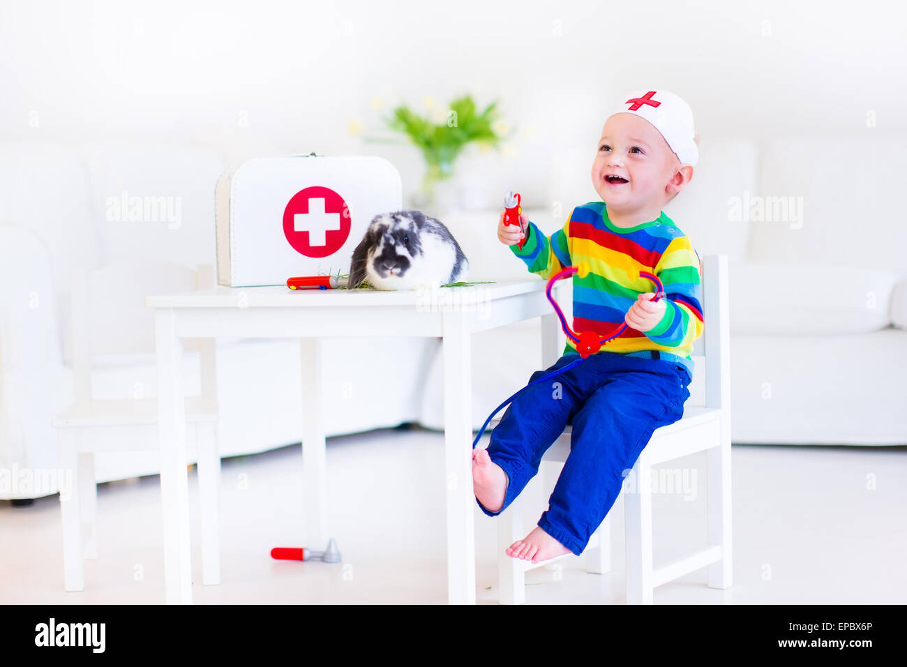 Carino Laughing baby boy riproduzione animale medico con il suo coniglio in pet utilizzando uno stetoscopio giocattolo in un bianco nursery room Foto Stock