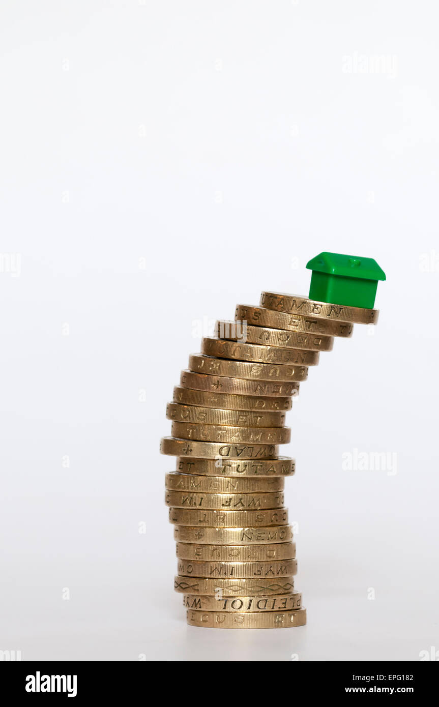Un piccolo modello di plastica green house bilanciato sulla sommità di una pila di inclinazione di libbra di monete. Finanziamento di alloggi concetto. Foto Stock