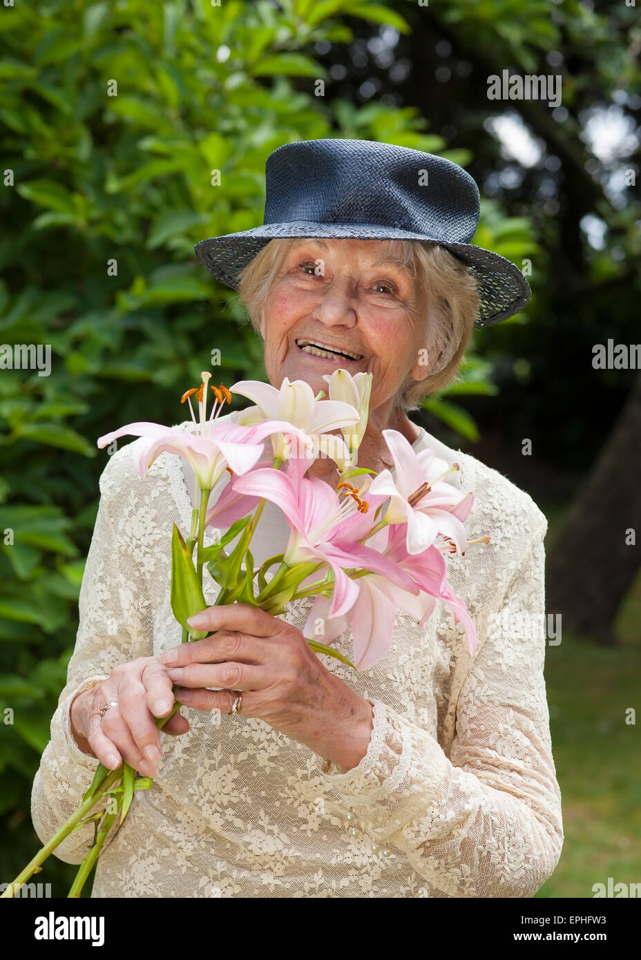 Felice signora anziana in un elegante abito di Lacy e hat all'aperto  permanente nel suo giardino con un bouquet di fiori freschi di gigli rosa  Foto stock - Alamy