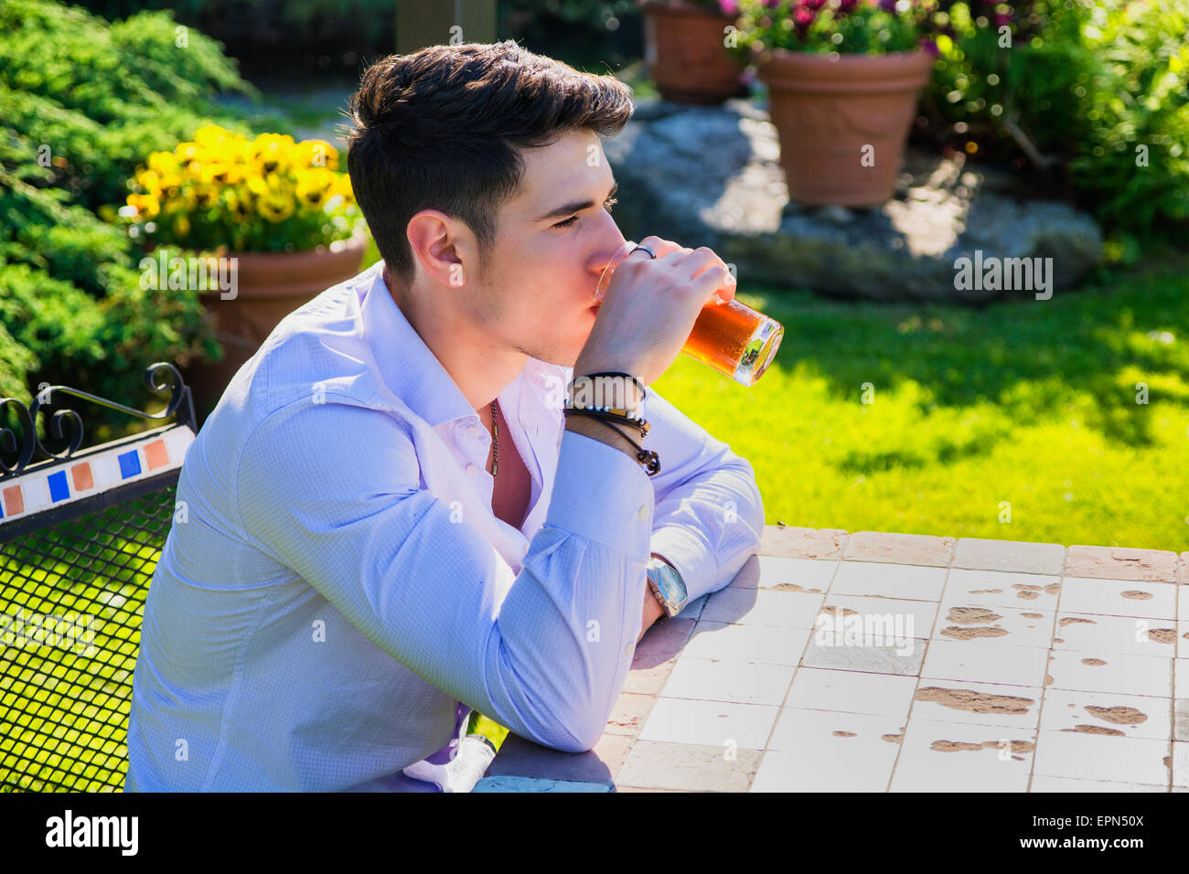 Bel giovane uomo seduto da solo al tavolo esterno in posizione di parcheggio o di natura, con bicchiere di bevanda di soda Foto Stock