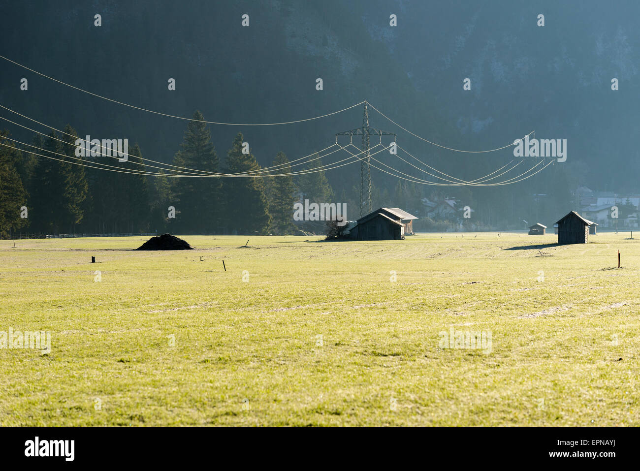 Una linea elettrica è un incrocio grean prato con alcune capanne di legno nella luce del mattino, Garmisch-Partenkirchen, Bavaria Foto Stock