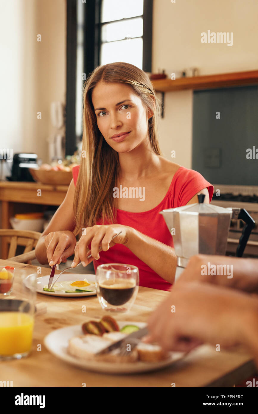 Ritratto di attraente giovane donna seduta al tavolo per la colazione e di mangiare guardando la fotocamera. Femmina caucasica avente Prima colazione in Foto Stock