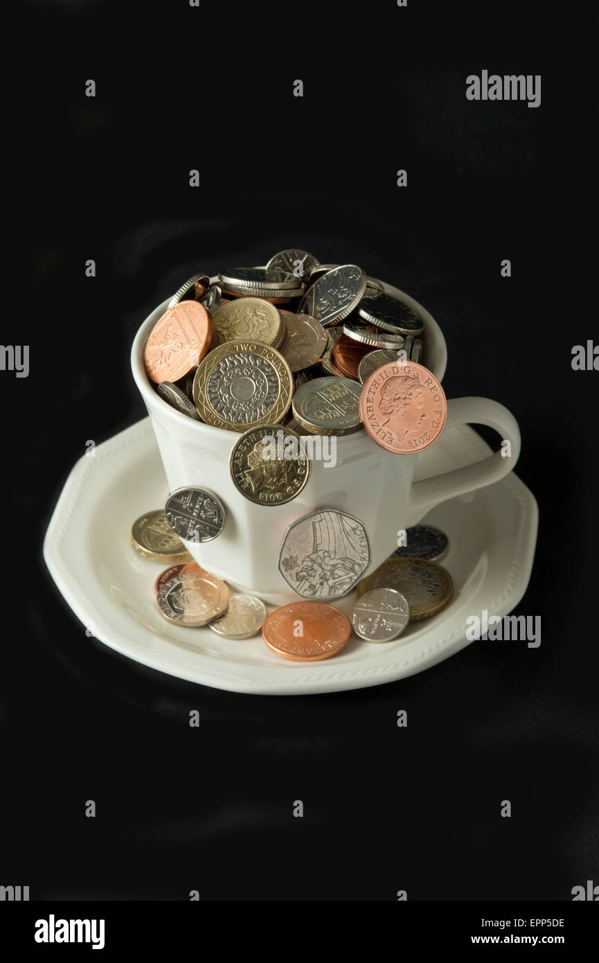 'My cup corre su un bicchiere di bianco e il piattino pieno di GBP British monete fuoriuscita.Un REGNO UNITO denaro economia finanza monete ricchezza Foto Stock