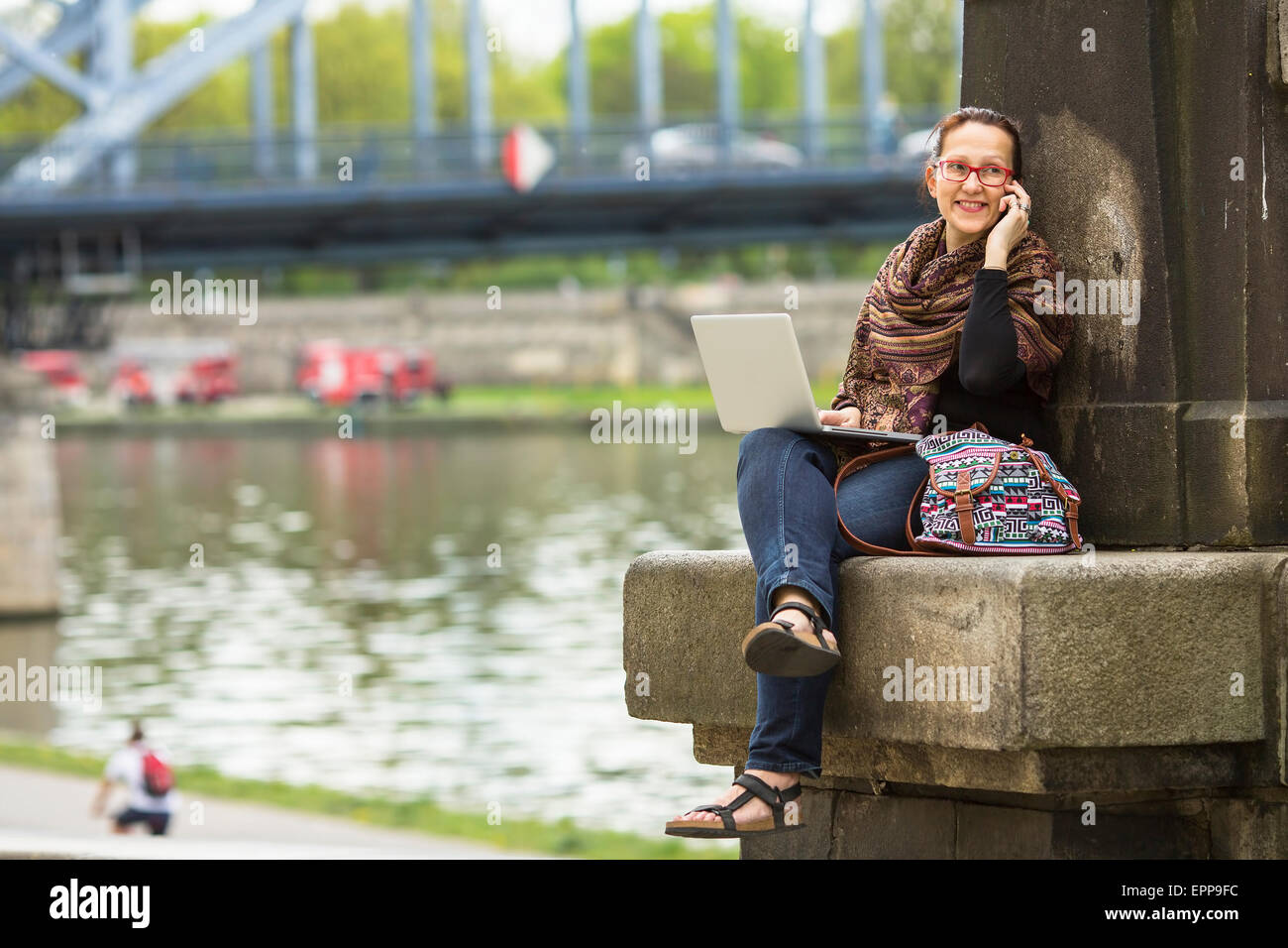 Giovane donna con il computer portatile seduti lungo gli argini del fiume nella città vecchia a parlare su un telefono cellulare. Foto Stock
