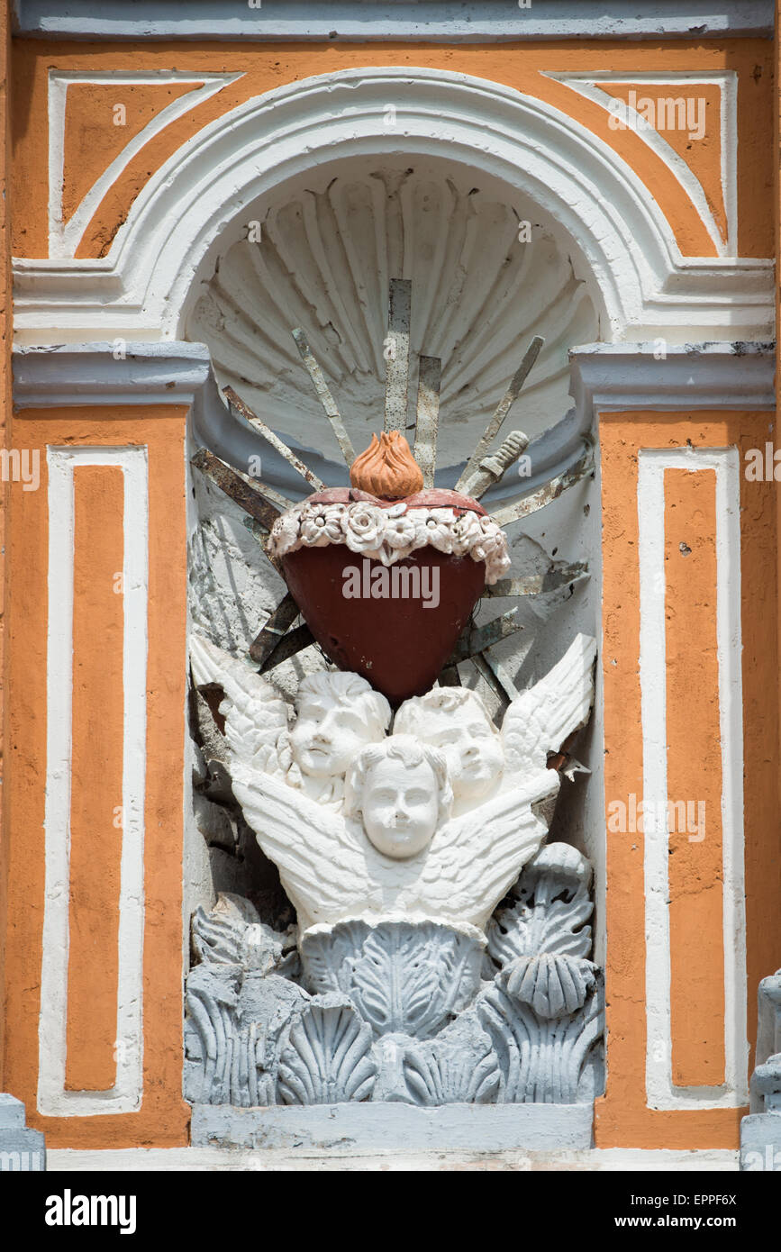 Iglesia de Xalteva Sacro cuore Scultura Granada Nicaragua // GRANADA, Nicaragua - Una scultura religiosa si trova in una piccola alcova sul muro esterno di Iglesia de Xalteva, una delle chiese storiche di Granada. La scultura in pietra presenta tre angeli alla base, che sostengono un simbolo del cuore sacro con una fiamma che emana dalla cima, che rappresenta l'amore ardente di Cristo per l'umanità. Questa iconografia cattolica del Sacro cuore con fiamma simboleggia l'amore divino, il sacrificio e il fervore spirituale. L'Iglesia de Xalteva risale al periodo coloniale e si erge come un importante punto di riferimento religioso a Gra Foto Stock