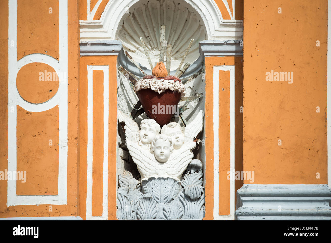 Iglesia de Xalteva Sacro cuore Scultura Granada Nicaragua // GRANADA, Nicaragua - Una scultura religiosa si trova in una piccola alcova sul muro esterno di Iglesia de Xalteva, una delle chiese storiche di Granada. La scultura in pietra presenta tre angeli alla base, che sostengono un simbolo del cuore sacro con una fiamma che emana dalla cima, che rappresenta l'amore ardente di Cristo per l'umanità. Questa iconografia cattolica del Sacro cuore con fiamma simboleggia l'amore divino, il sacrificio e il fervore spirituale. L'Iglesia de Xalteva risale al periodo coloniale e si erge come un importante punto di riferimento religioso a Gra Foto Stock
