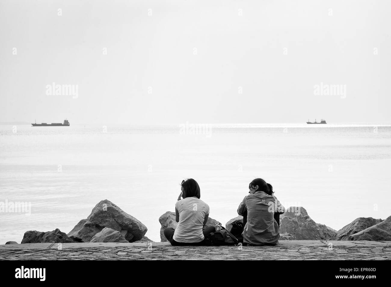 Due ragazze seduti sulla passeggiata lungo il viale miramare guardando le navi passare da nel golfo di Trieste, Italia. Foto Stock