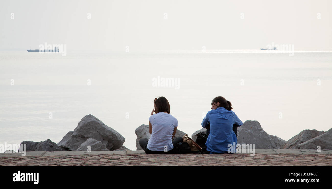 Due ragazze seduti sulla passeggiata lungo il viale miramare guardando le navi passare da nel golfo di Trieste, Italia. Foto Stock