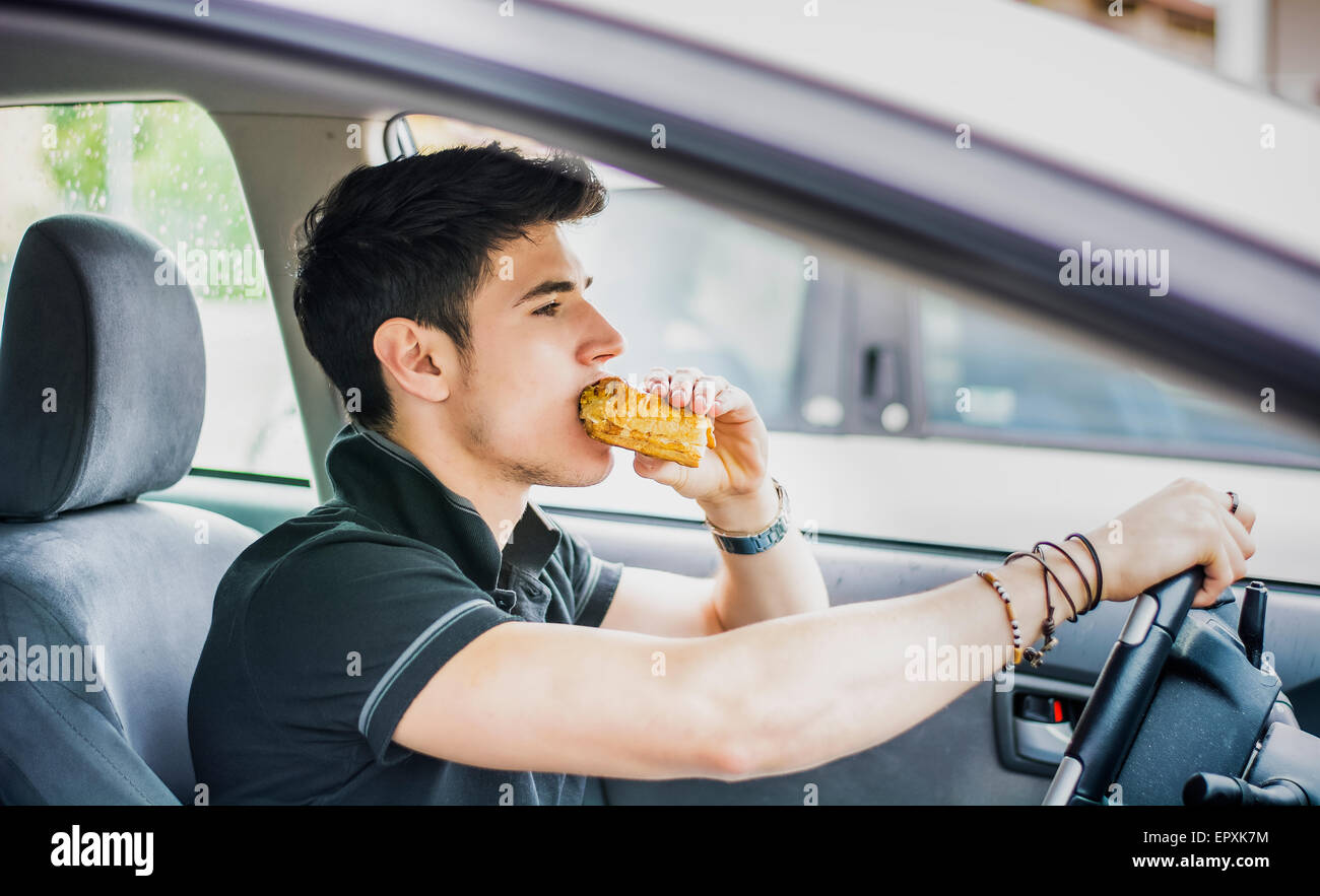 Giovane uomo bello alla guida della sua auto mentre mangiando cibo nel traffico Foto Stock