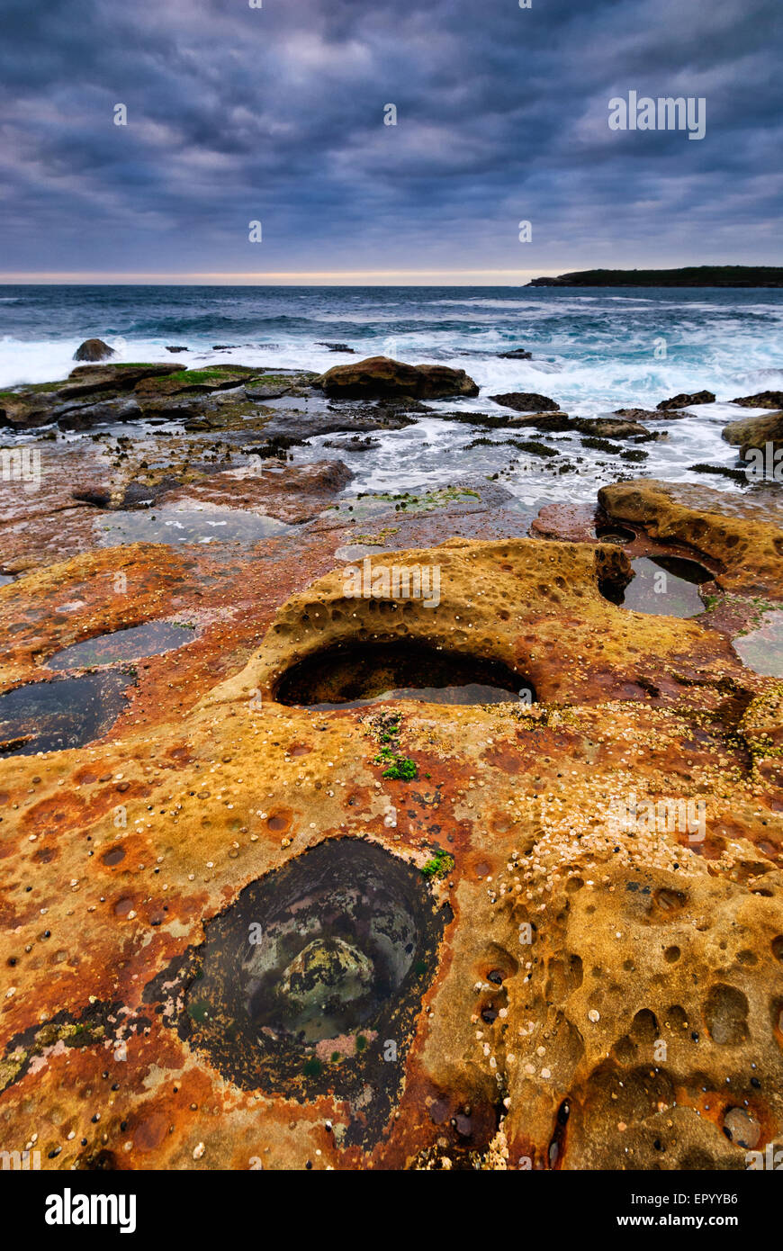 Sunrise seascape con rocce di colore arancione e oceano piscine e conchiglie di mare con nuvoloso cielo tempestoso e scogliere distanti Foto Stock