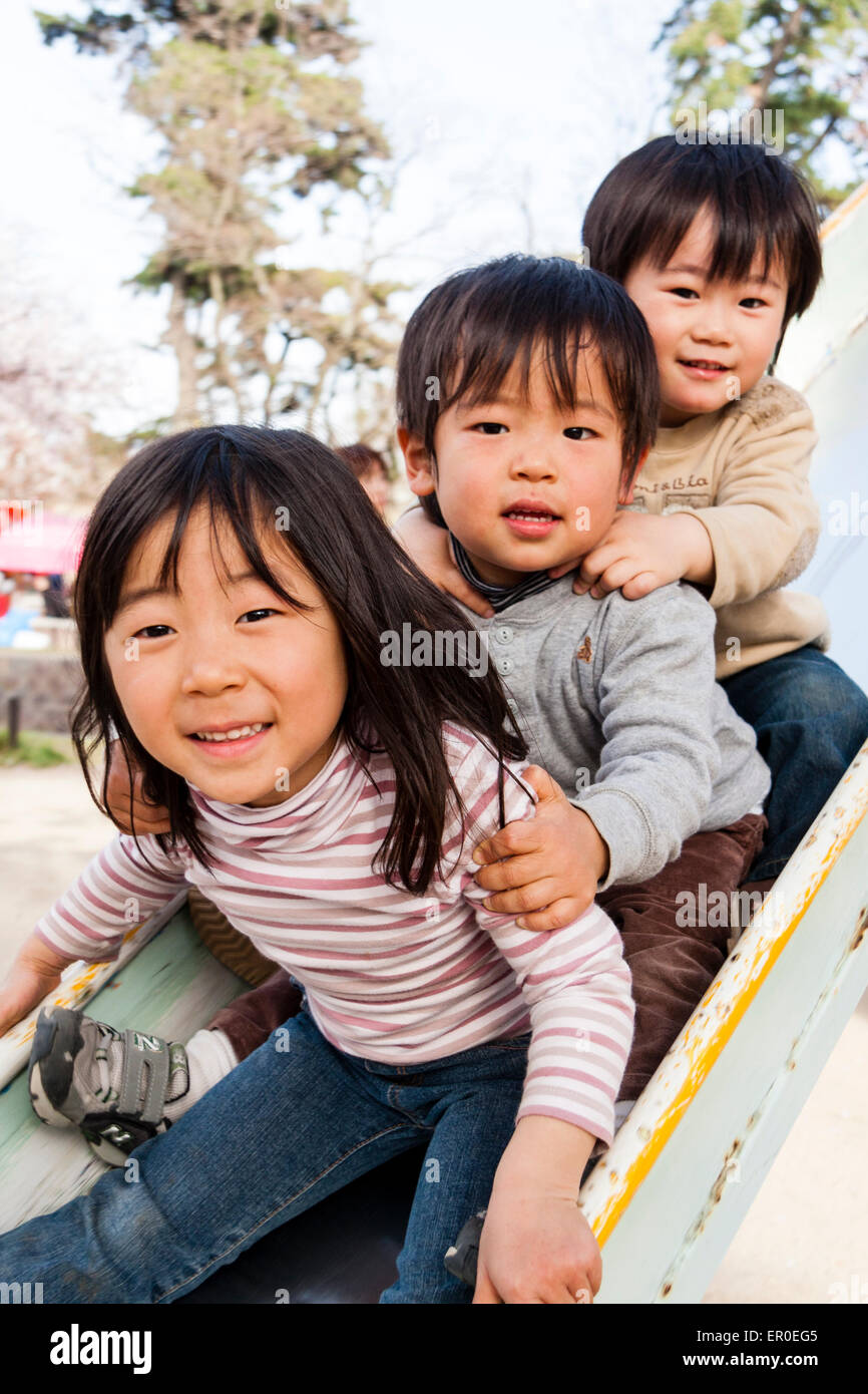Tre bambini giapponesi, ragazza, due ragazzi, 4-5 anni, seduti su uno scivolo che si tiene sulle spalle, guardando lo spettatore e sorridendo. Foto Stock