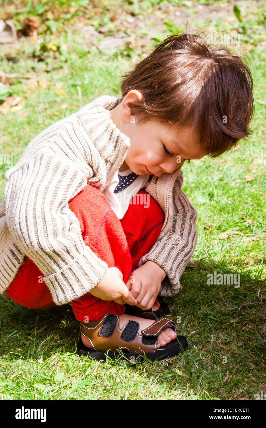 Bambino piccolo, ragazzo, 3-4 anni, accovacciato su erba in giardino per  allacciare le cinghie di sandalo. Indossa un cardigan spesso e pantaloni  rossi Foto stock - Alamy