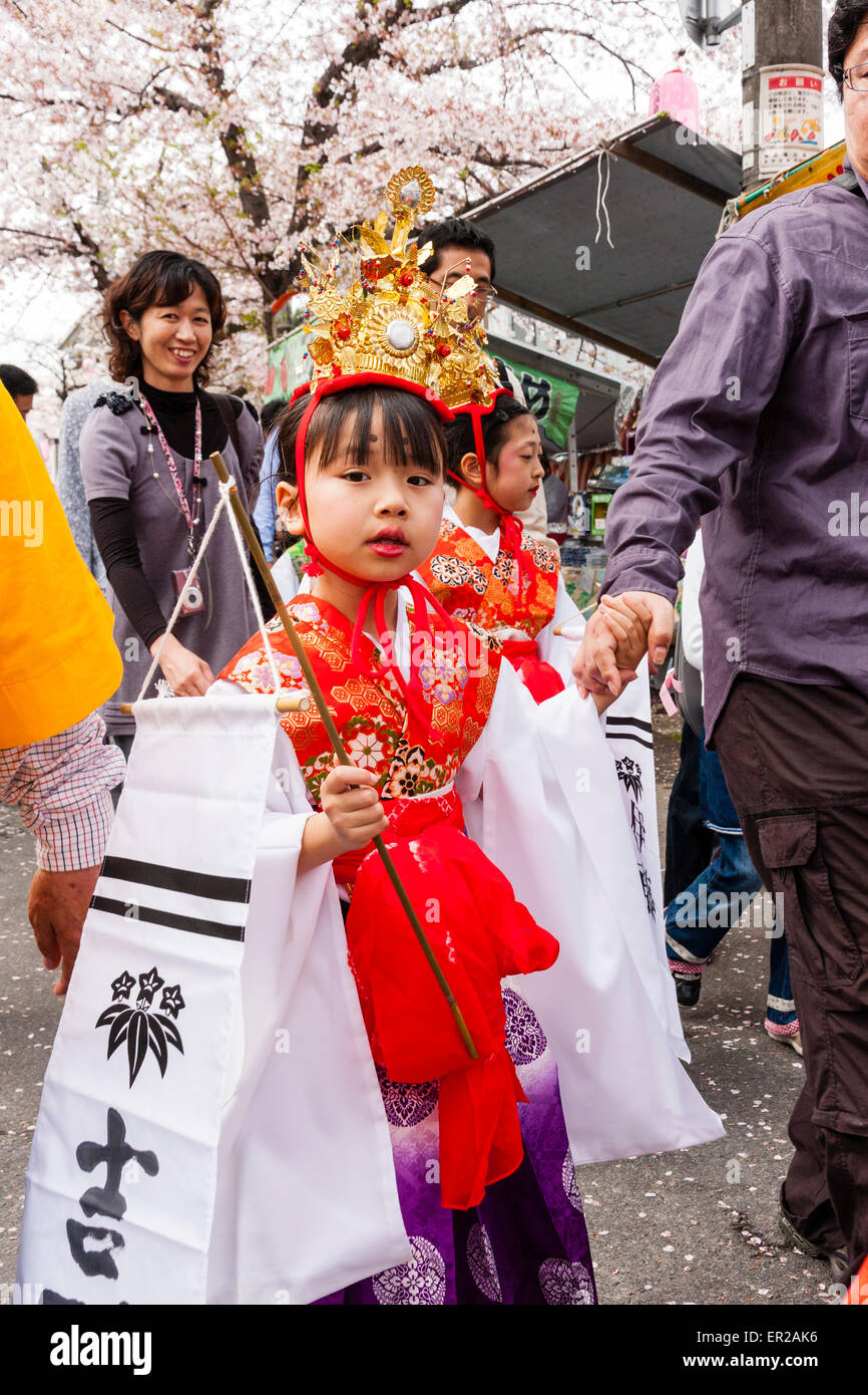 Bambino, ragazza, che indossa una giacca rossa con maniche di seta bianca e una corona d'oro che cammina nella sfilata primaverile Genji a Tada, Giappone. Contatto visivo. Foto Stock