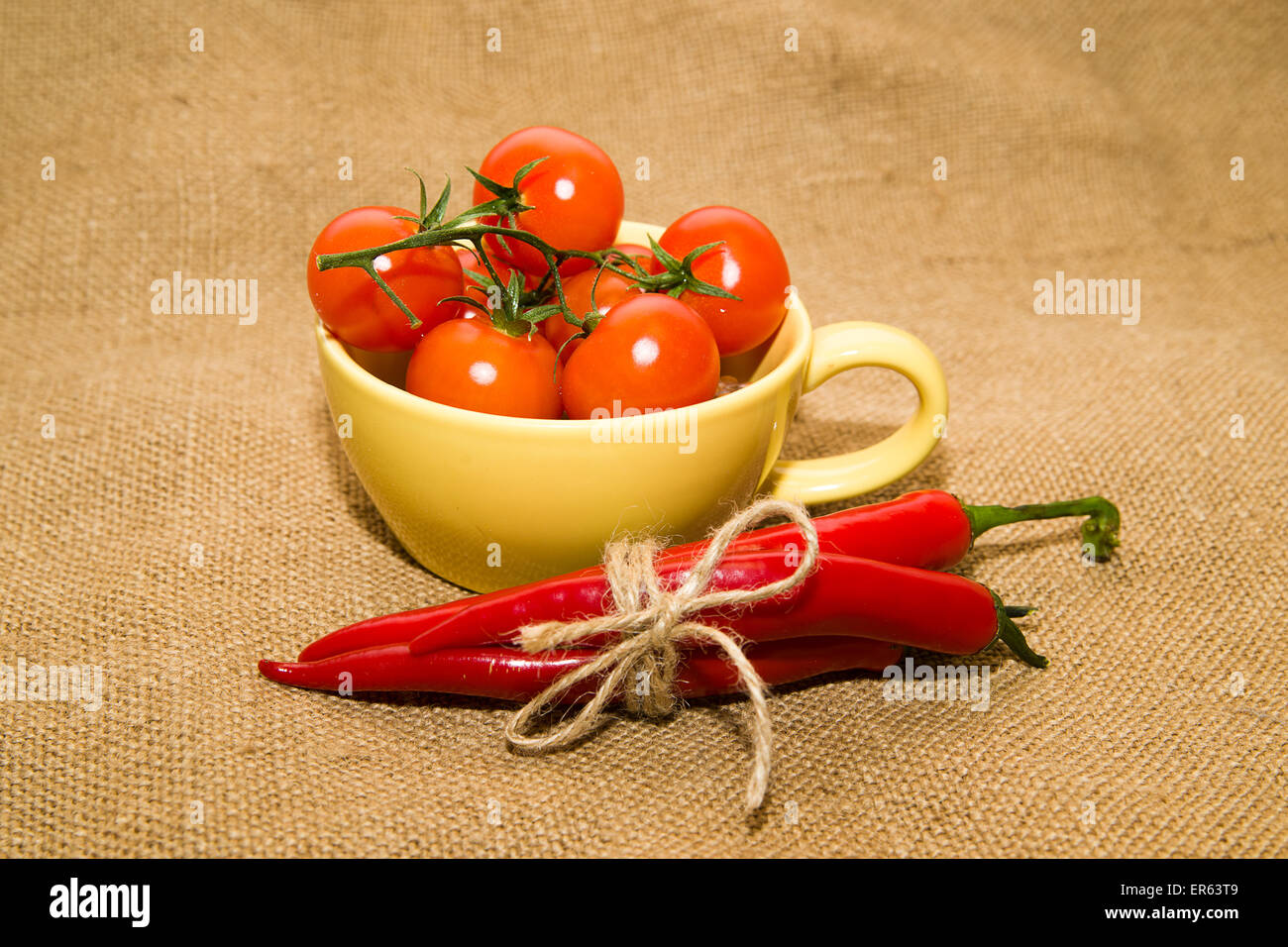 Pomodori ciliegia in una tazza di colore giallo e i peperoncini legato con una corda sul panno vecchio Foto Stock