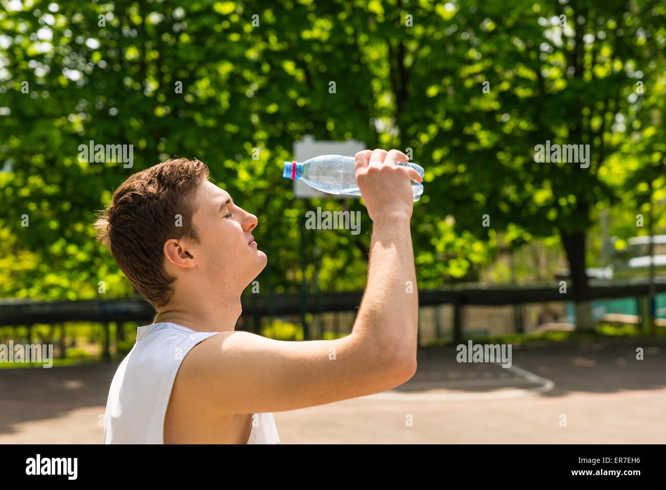 Testa e spalle vista del giovane uomo atletico circa per versare l'acqua dalla bombola sul viso, prendendo una pausa di ristoro e idratazione sul campo da basket. Foto Stock