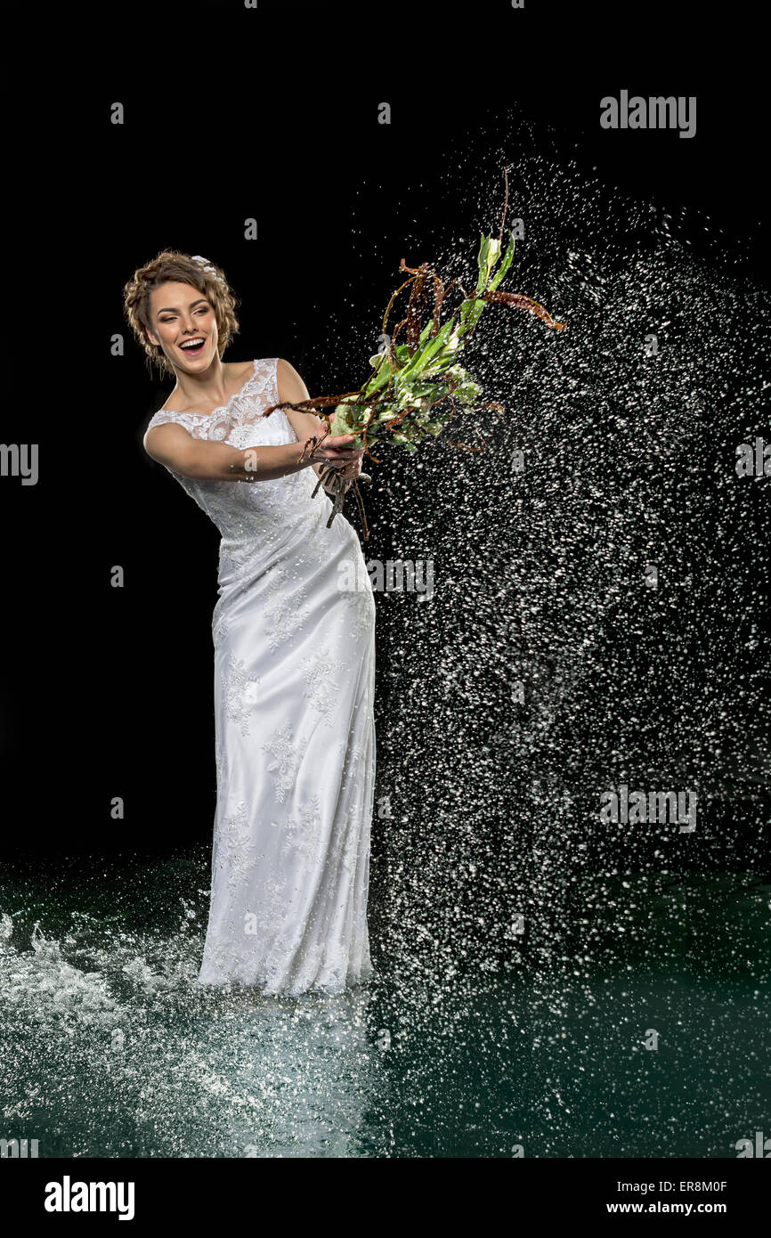 Eccitato giovane sposa gli spruzzi di acqua con il mazzo di fiori su sfondo nero Foto Stock