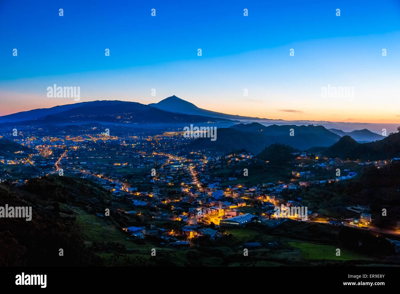 Città o comune con illuminazione dopo il tramonto in serata in montagna con cielo blu e il vulcano Teide sullo sfondo in Tenerife Foto Stock