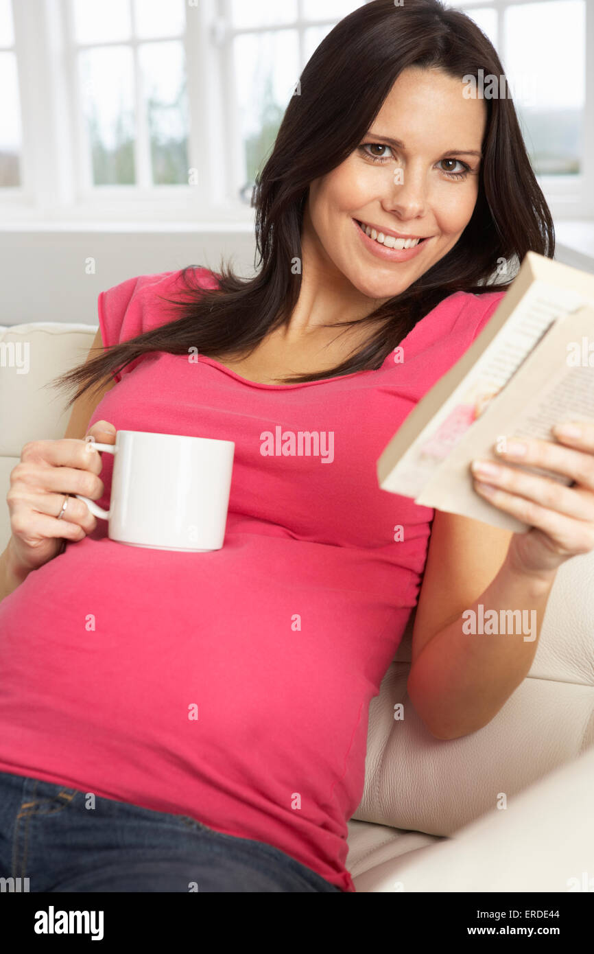 Donna incinta di bere una bevanda calda e libro di lettura a casa Foto Stock