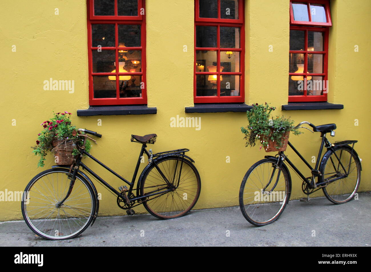 Due biciclette antiquate con cestini di fiori al manubrio, appoggiate contro una parete di stucco rosso e giallo brillante, un'immagine nostalgica. Foto Stock
