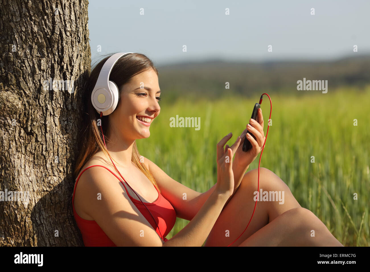 Ragazza ascoltando la musica e il download di brani seduto in un campo verde che indossa una maglietta rossa Foto Stock