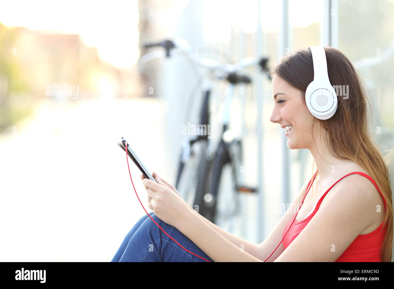 Donna ascoltando la musica da una tavoletta di seduta sul pavimento in un parco con una bicicletta in background Foto Stock