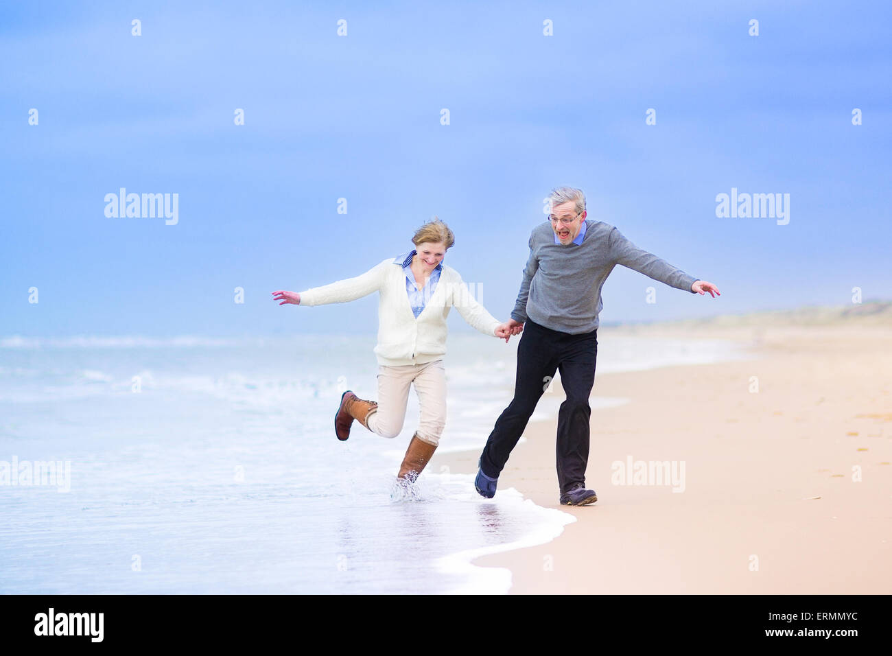 Felice Coppia di mezza età, bella donna attiva e laughing man in esecuzione su una spiaggia saltando lontano dalle onde del mare del Nord, Foto Stock