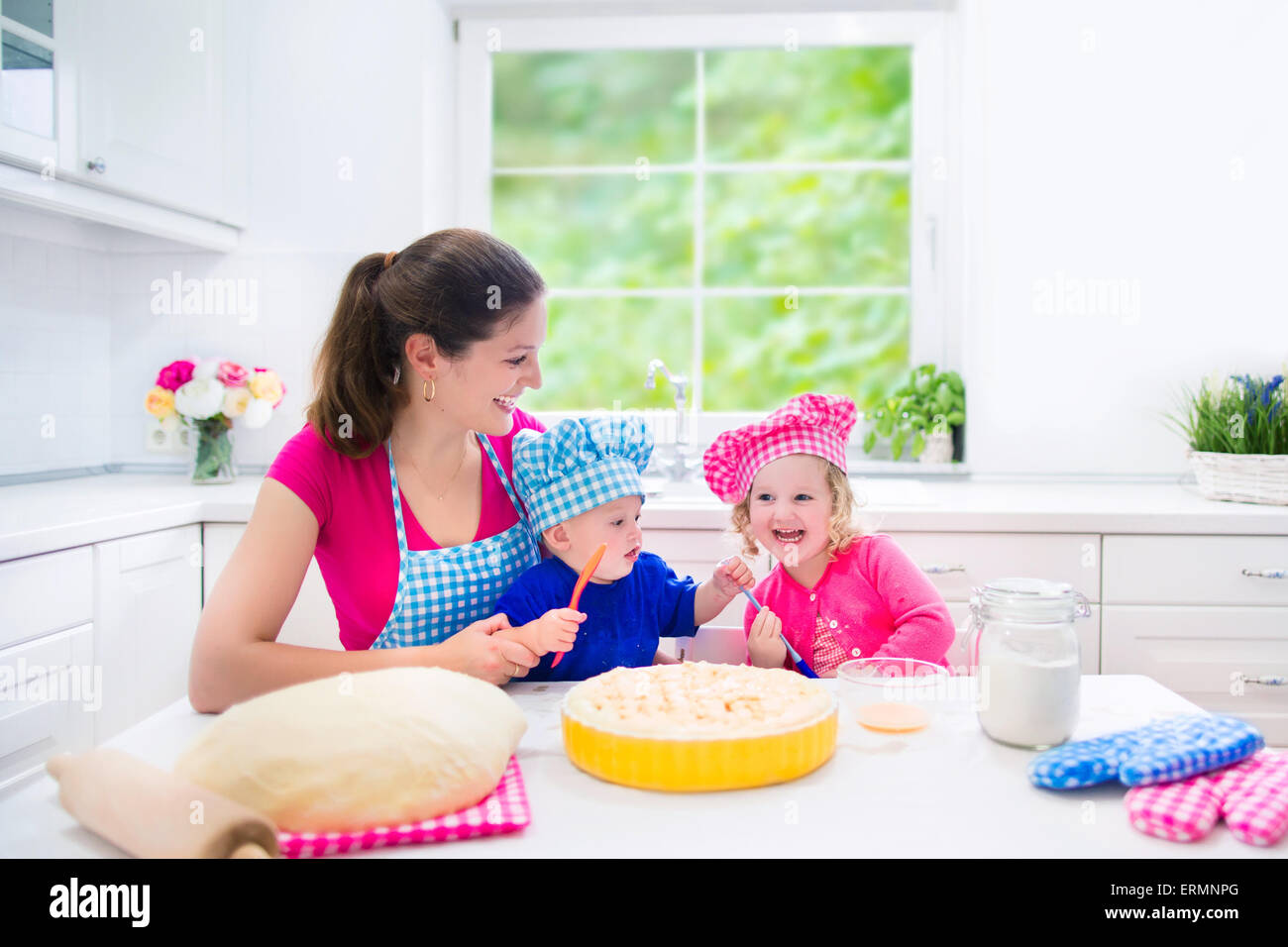 Felice giovane madre e i suoi bambini che indossano i colori rosa e blu  chef cappelli cottura di una torta insieme in una soleggiata bianco cucina  con grande finestra Foto stock - Alamy