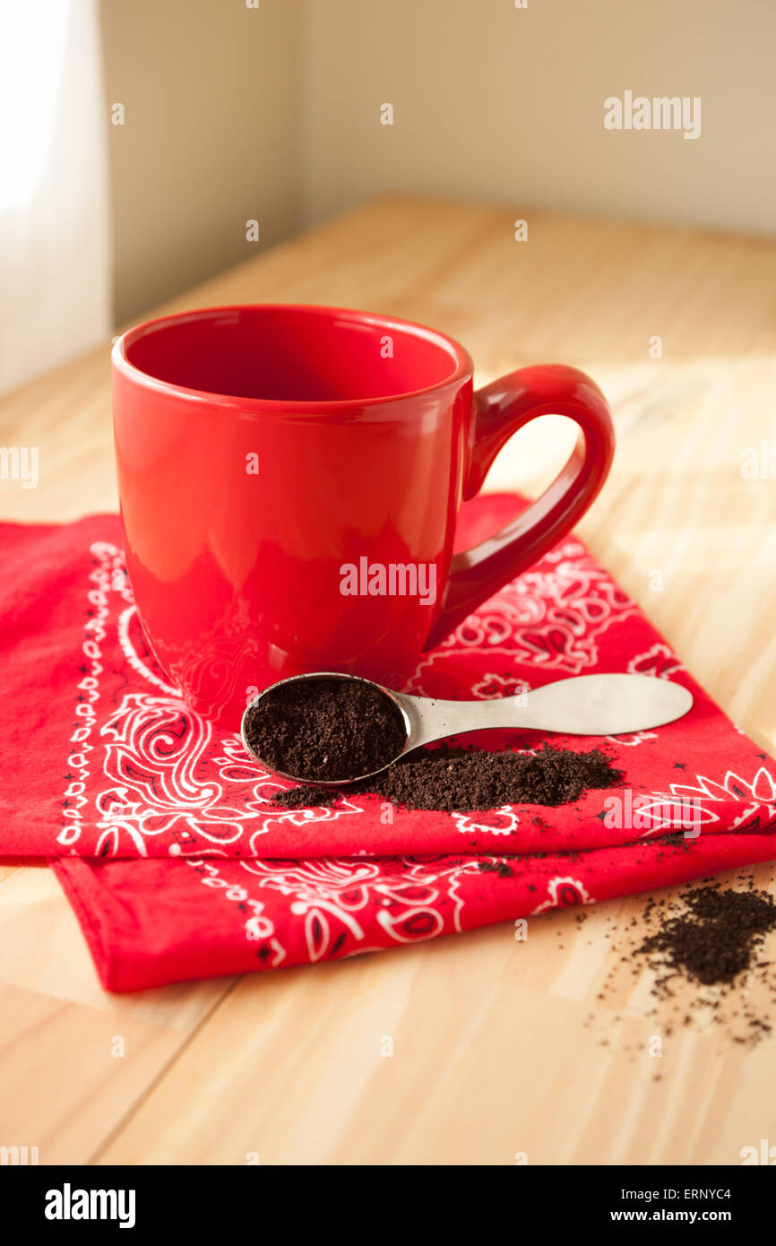 Un rosso tazza da caffè si trova sulla cima di un rosso bandana igienico. Un cucchiaio di caffè macinato viene versato intorno e si siede accanto alla tazza. Foto Stock