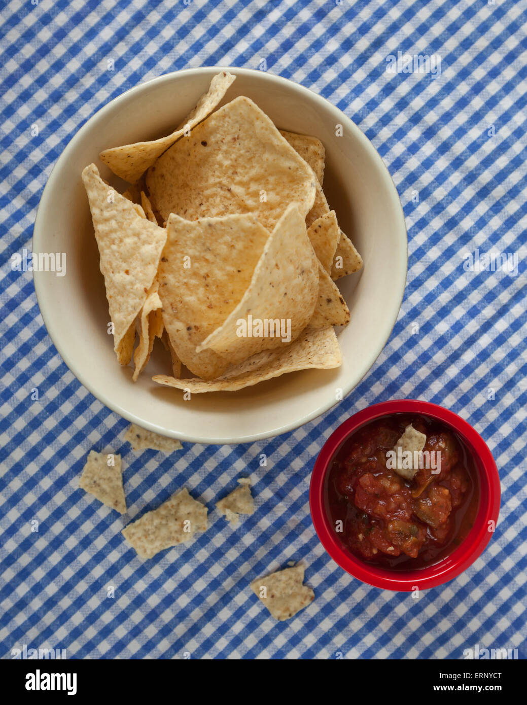 Una ciotola di corn tortilla chips sedersi accanto a un recipiente rosso di salsa su un blu e bianco tovaglia a scacchi. Pezzi di tortilla sono b Foto Stock