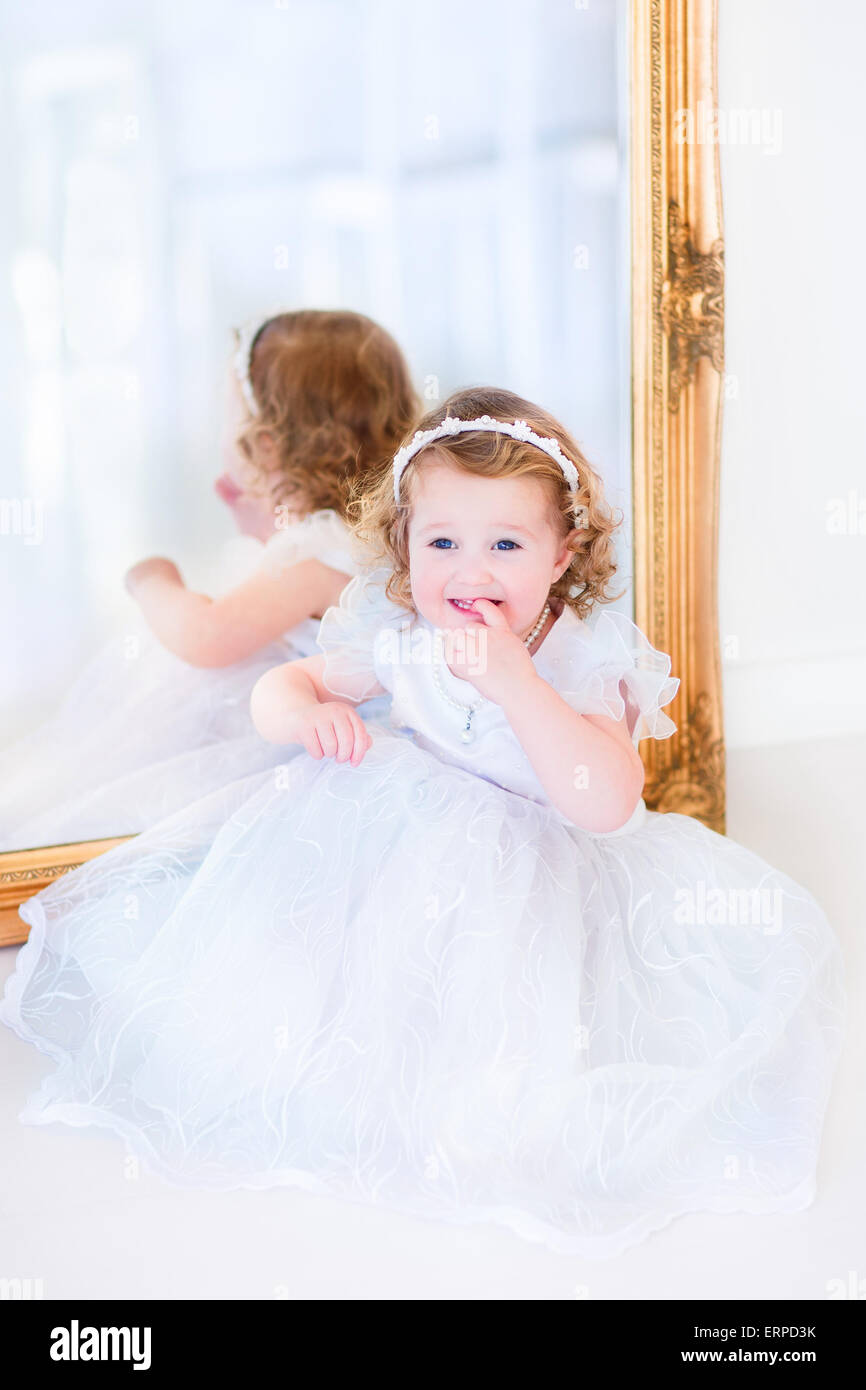Ricci dolce bambina con grande bella occhi indossando un bianco damigella abito seduta a una grande finestra Riproduzione princess Foto Stock