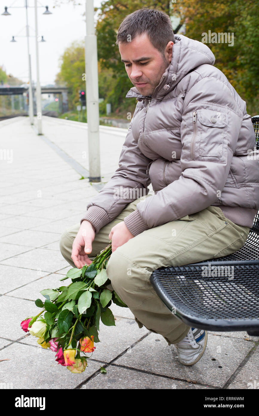 Uomo bello in attesa alla stazione ferroviaria con rose e guardando deluso Foto Stock