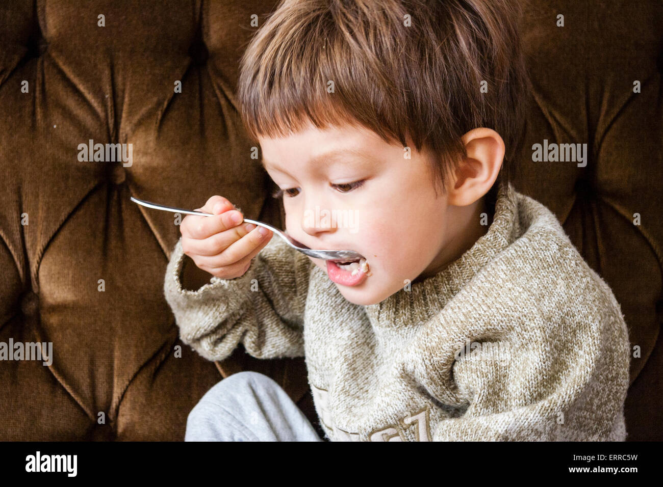 Interno, primo piano di un bambino caucasico, ragazzo, di 3-4 anni, con un grosso ponticello e seduto sul divano marrone mentre si mangia cereali con un cucchiaio d'argento. Foto Stock