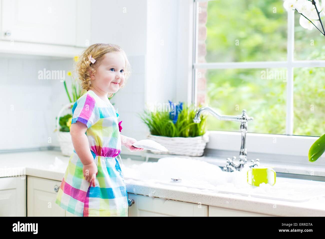 Carino curly toddler ragazza in un abito colorato lavaggio piatti, la pulizia con una spugna e giocare con la schiuma nel lavandino Foto Stock