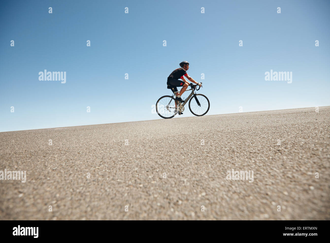 Basso angolo immagine di un ciclista maschio a cavallo su una strada piana contro il cielo blu. Uomo in bicicletta in salita su strada aperta. Foto Stock