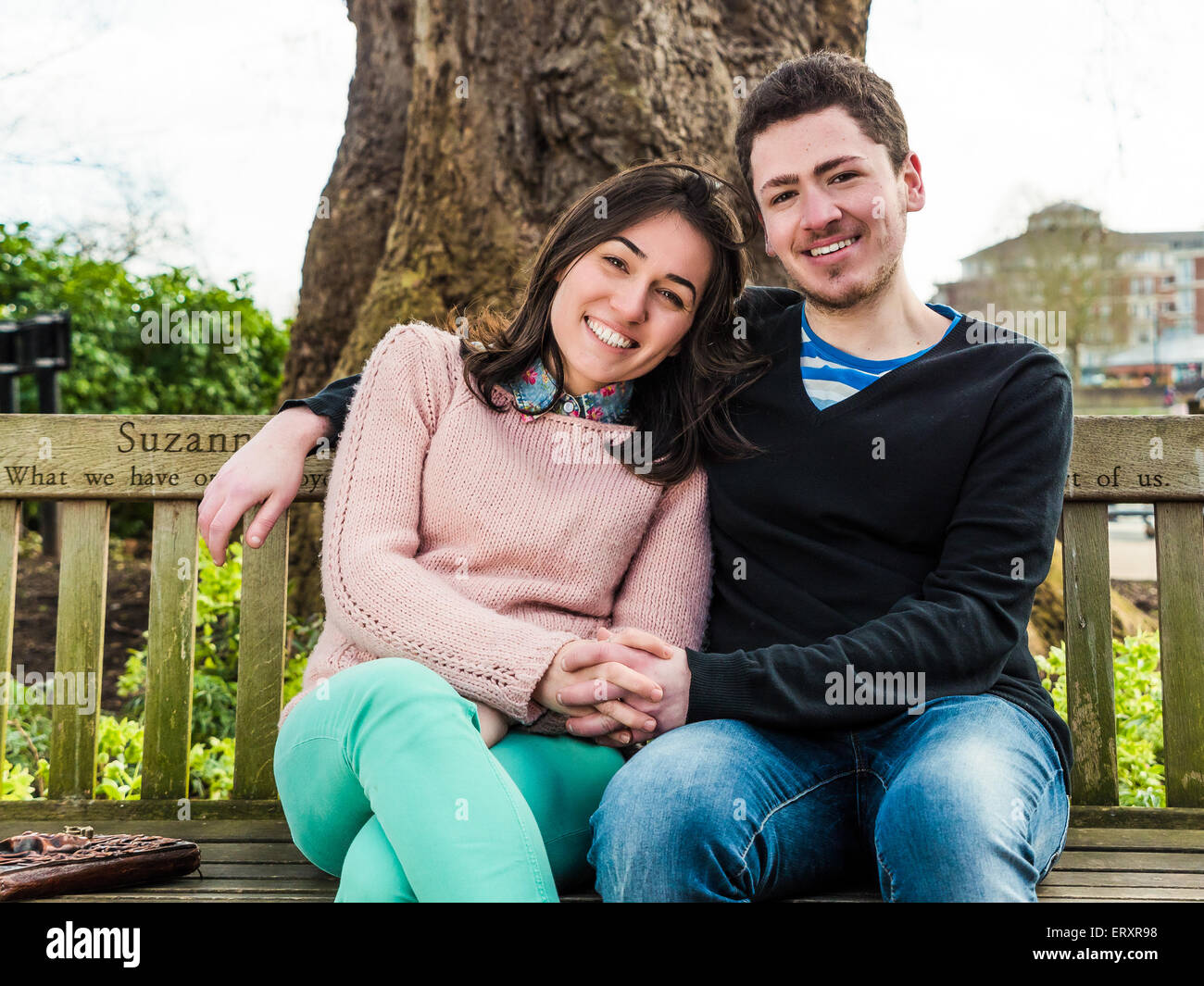 Giovane coppia eterosessuale seduta su una panchina del Parco godendo di una splendida giornata di sole e sorridente guardando la telecamera Foto Stock