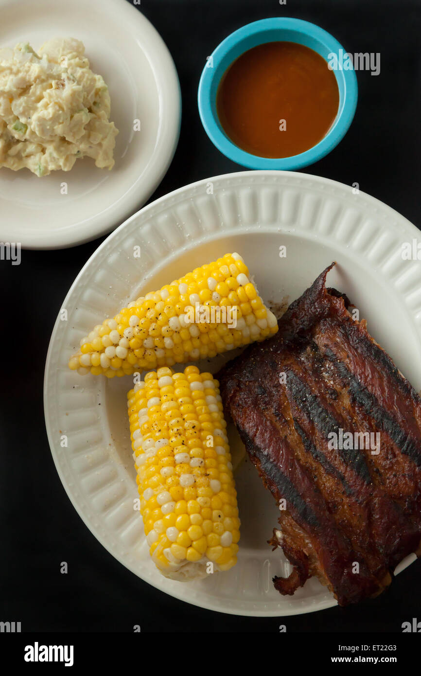Costolette di maiale su di una piastra con sulla pannocchia di mais, insalata di patate, tè freddo e salsa barbecue Foto Stock