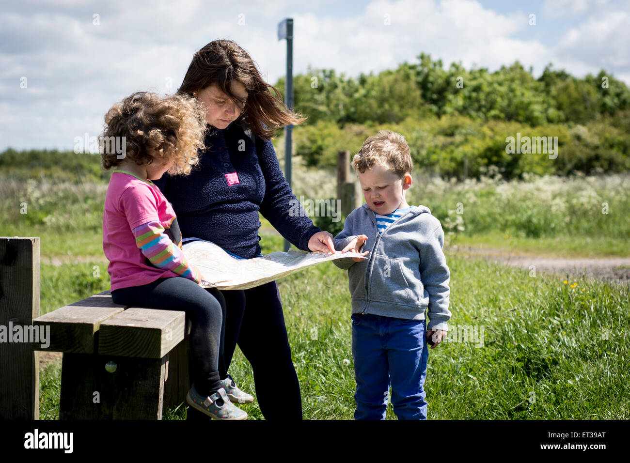Madre e bambini ragazza ragazzo famiglia seguente una cartina passeggiate in campagna con il sole splendente e nuvole nel cielo Foto Stock
