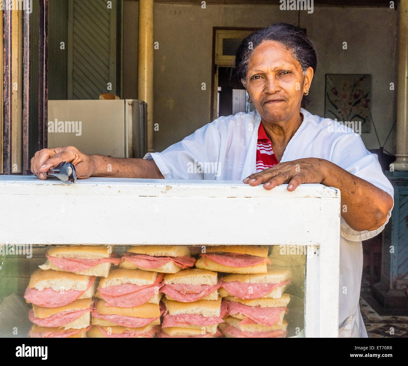 Un vecchio afro-cubane sandwich femmina venditore si appoggia sul suo caso di visualizzazione azienda panini e altri alimenti mantenendo le tenaglie. Foto Stock