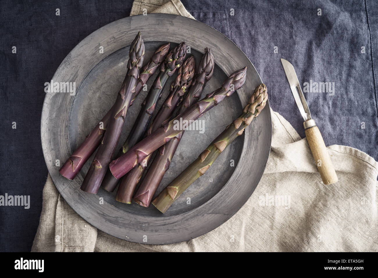 Freschi asparagi viola sul peltro vintage piastra con peeling vintage coltello sulla biancheria scura Foto Stock