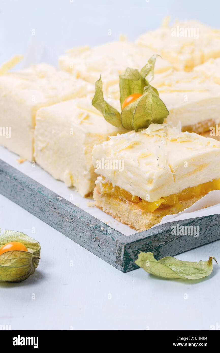 In casa fette di torta con mousse cremosa e frutti tropicali di mango e physalis servita nel vassoio in legno su fondo azzurro Foto Stock