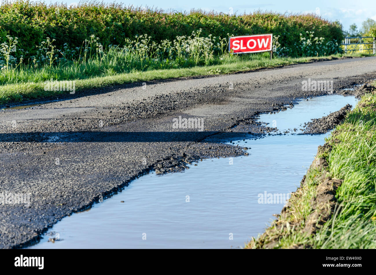 Riempito di acqua buca sulla strada di un paese con un 'Slow' firmare in background Foto Stock