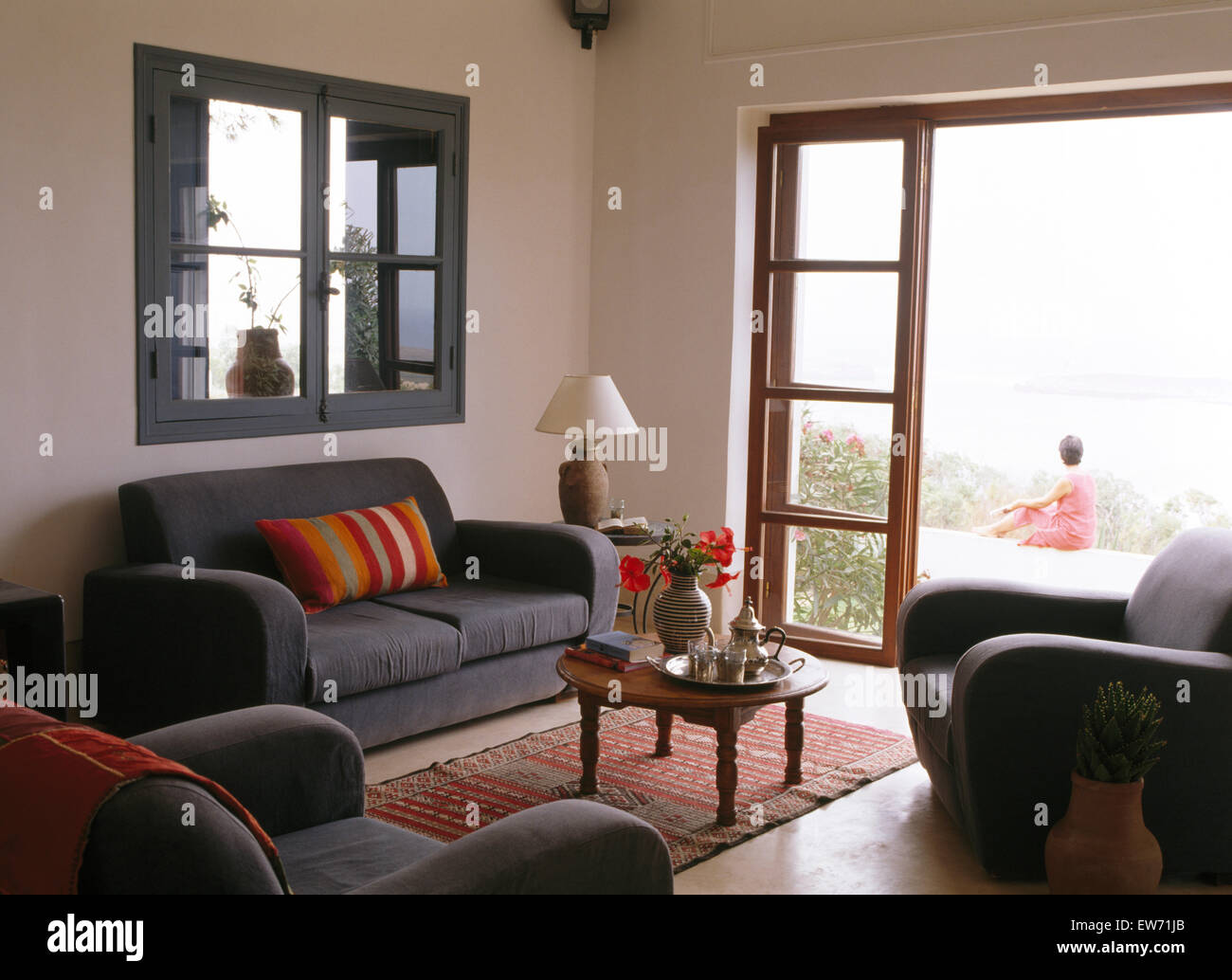 Divano grigio sotto finestra interno marocchino in salotto con vista attraverso le porte di vetro della donna seduta sul patio Foto Stock