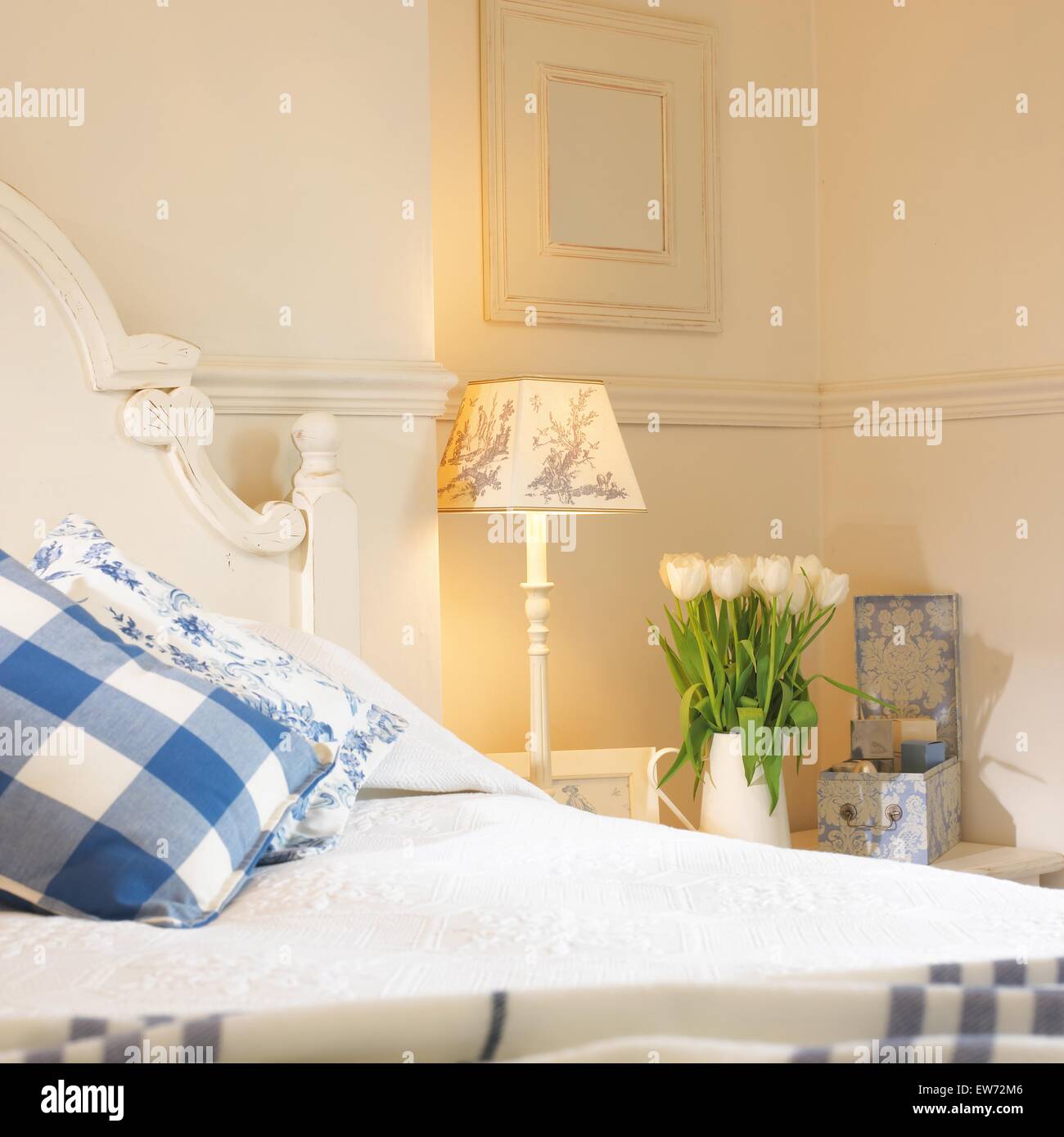 Blue+bianco a scacchi cuscini sul letto dipinte di bianco camera da letto con una lampada accesa e vaso di tulipani bianco sul comodino Foto Stock