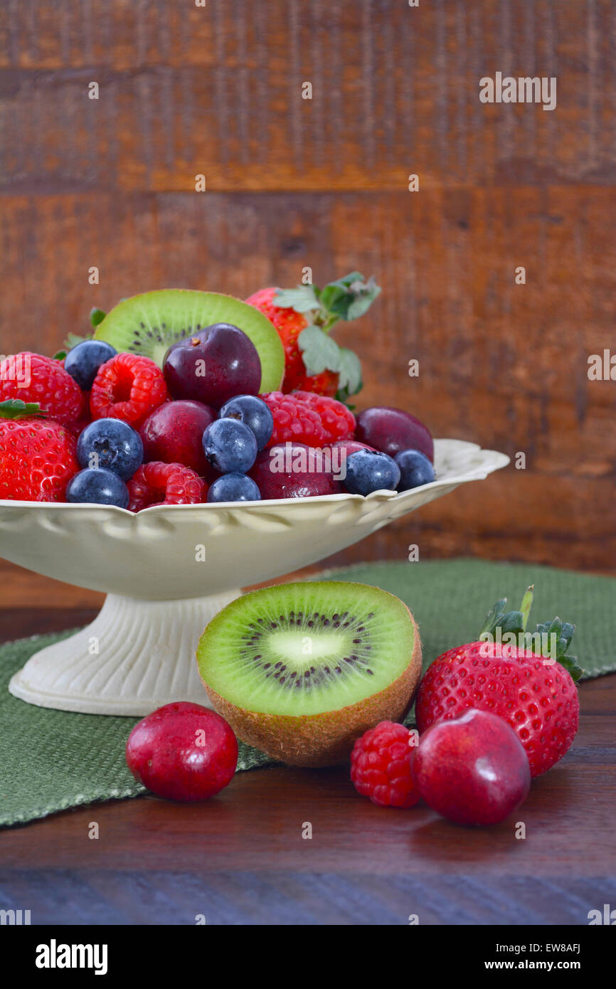 Frutta estiva compresi lamponi, fragole, ciliegie, mirtilli e kiwi in una ciotola vintage con rigenerati woo scuro Foto Stock