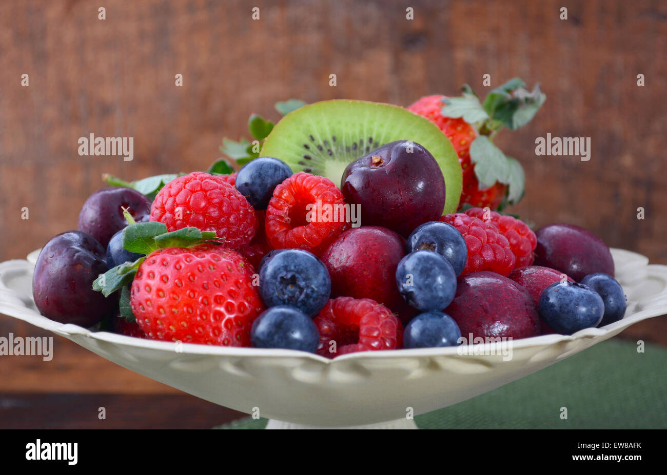 Frutta estiva compresi lamponi, fragole, ciliegie, mirtilli e kiwi in una ciotola vintage con rigenerati woo scuro Foto Stock