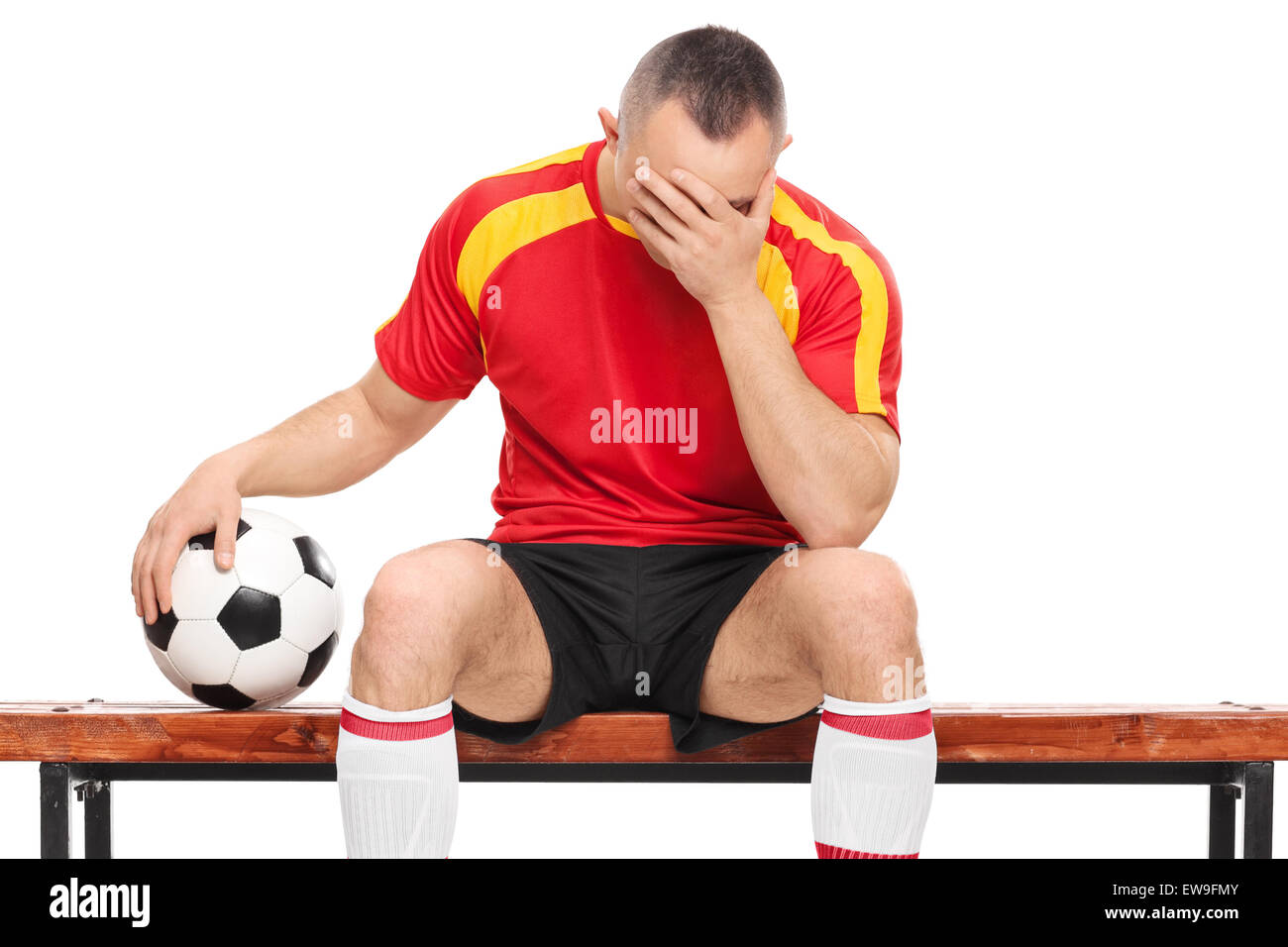 Preoccupato per il giocatore di football seduto su una panca in legno, tenendo una palla e nasconde la sua faccia con una sola mano isolato su bianco Foto Stock