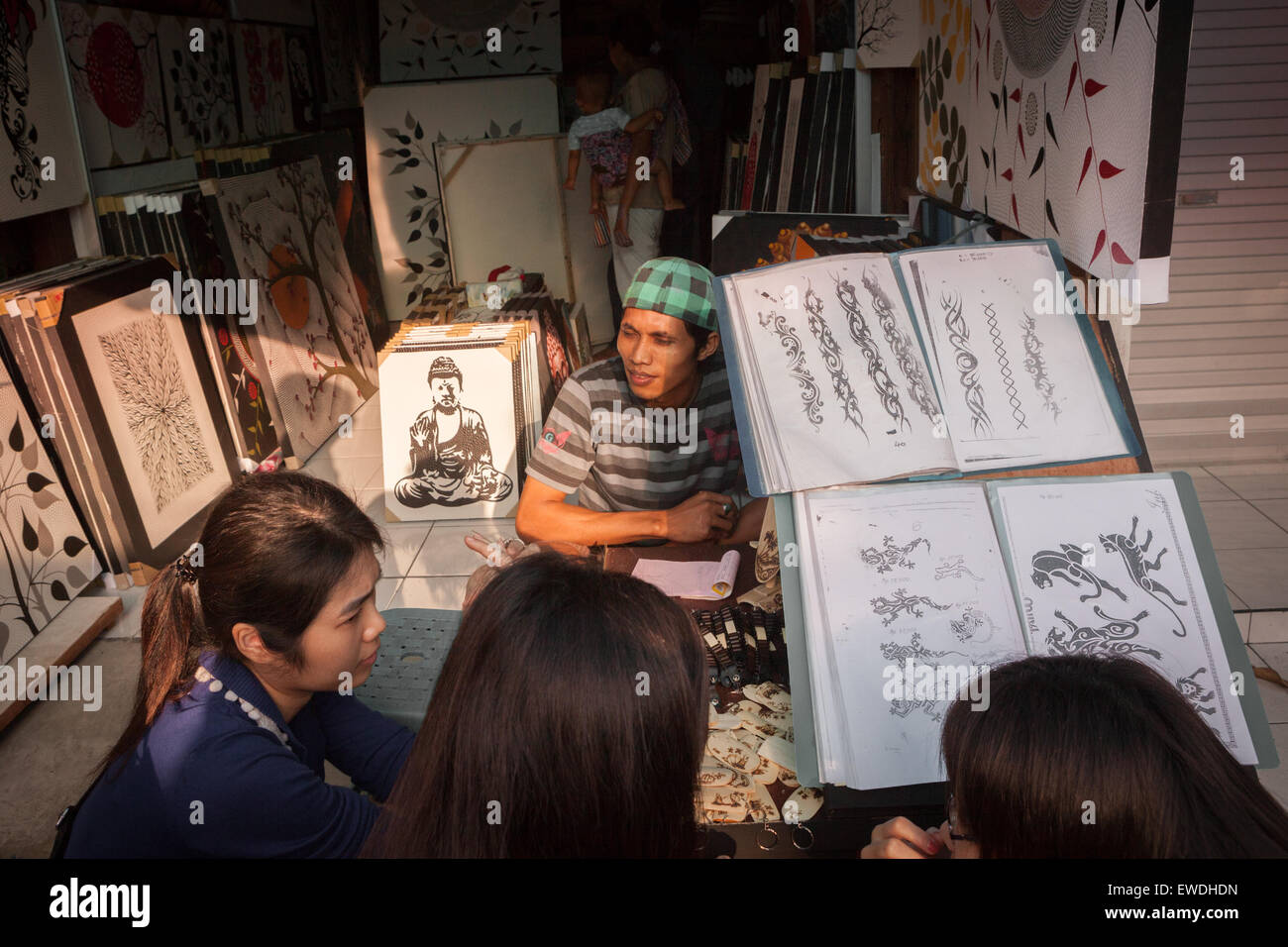 Un acquirente aspetta come un gruppo di turisti donne stanno discutendo le opzioni mentre accovacciandosi davanti ad un grande libro di esposizione di disegno del tatuaggio a Bali. Foto Stock