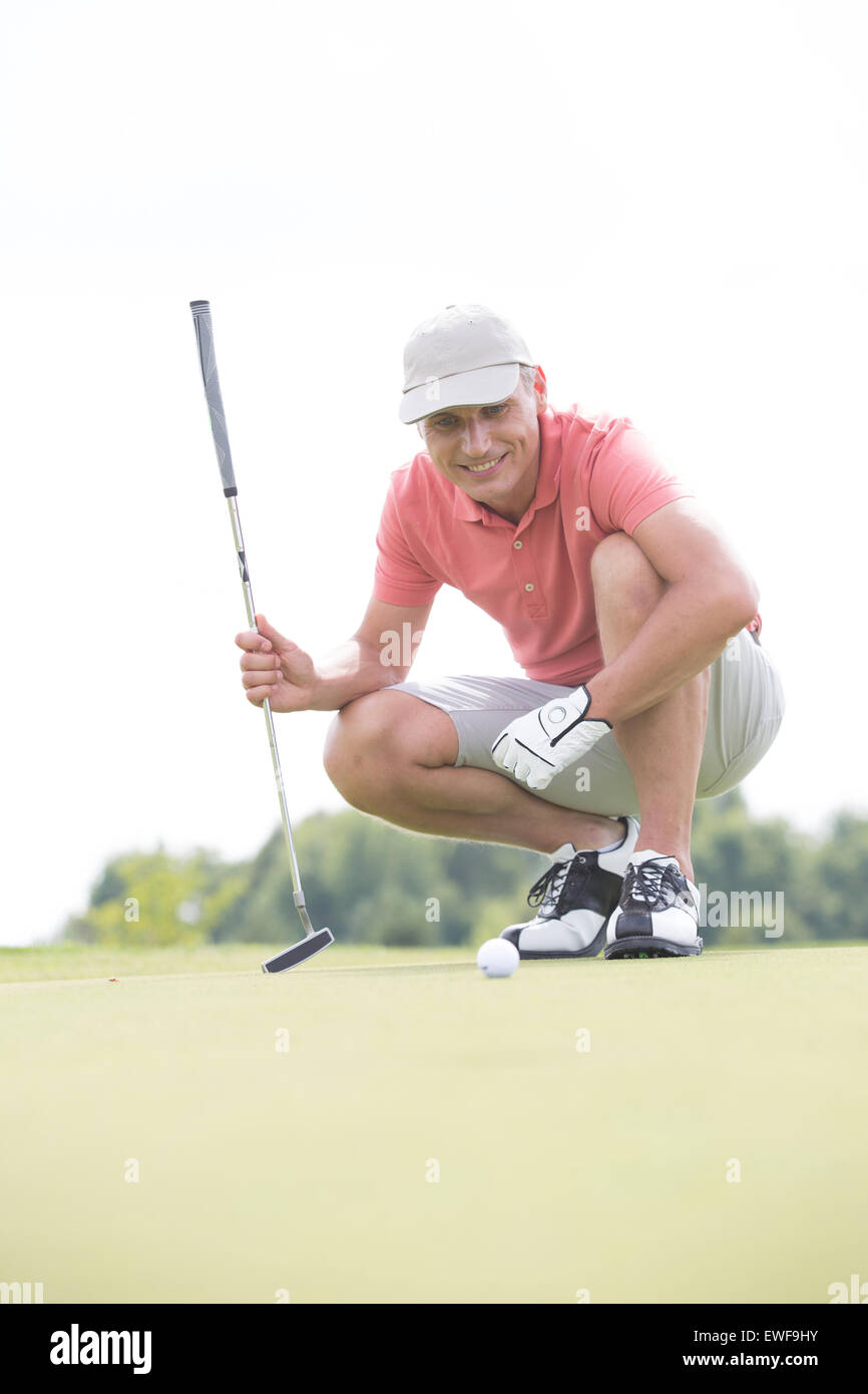 Sorridente uomo di mezza età guardando a sfera mentre accovacciato sul campo da golf Foto Stock